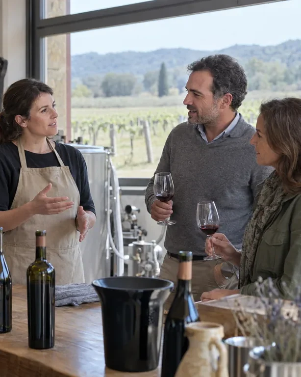 Un'enologa conversa con due visitatori che tengono il calice durante una degustazione, con il vigneto oltre la vetrata